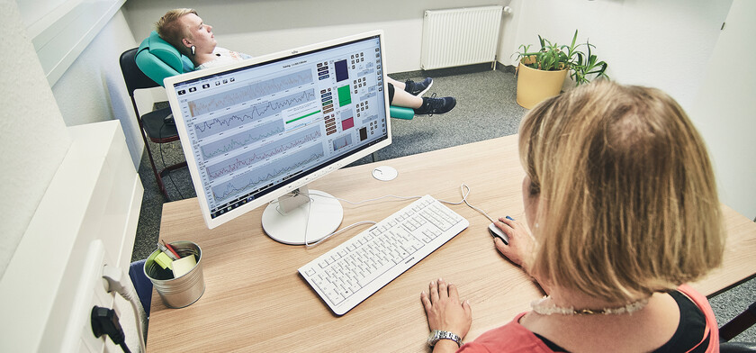 Ein Auszubildender liegt auf einer Liege im Büro für Neurofeedback. Eine Frau sitzt am Computer.